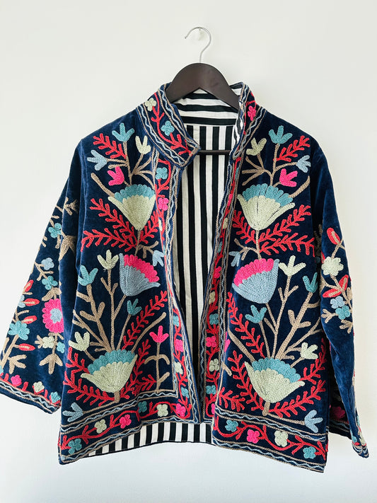 Colorful royal blue embroidered velvet jacket on a hanger against a white wall. Luxurious holiday outfit made by artisans in India for Ornate Handicrafts