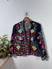 Colorful royal blue embroidered velvet jacket on a hanger against a white wall. Luxurious holiday outfit made by artisans in India for Ornate Handicrafts