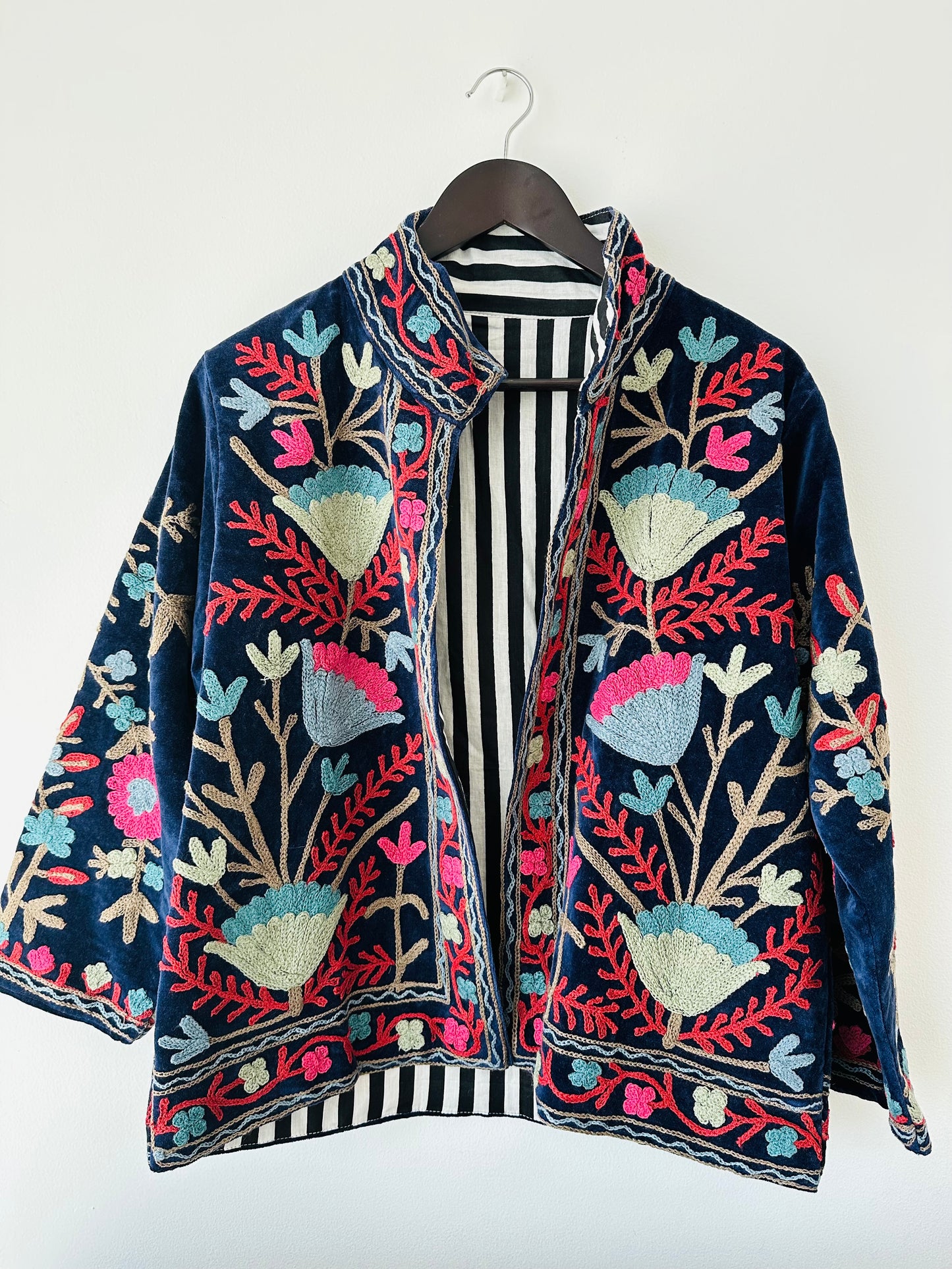 Colorful royal blue embroidered velvet jacket on a hanger against a white wall. Luxurious holiday outfit made by artisans in India for Ornate Handicrafts