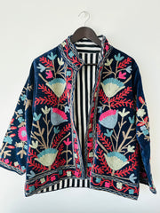 Colorful royal blue embroidered velvet jacket on a hanger against a white wall. Luxurious holiday outfit made by artisans in India for Ornate Handicrafts