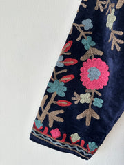 Colorful royal blue embroidered velvet jacket on a hanger against a white wall. Luxurious holiday outfit made by artisans in India for Ornate Handicrafts