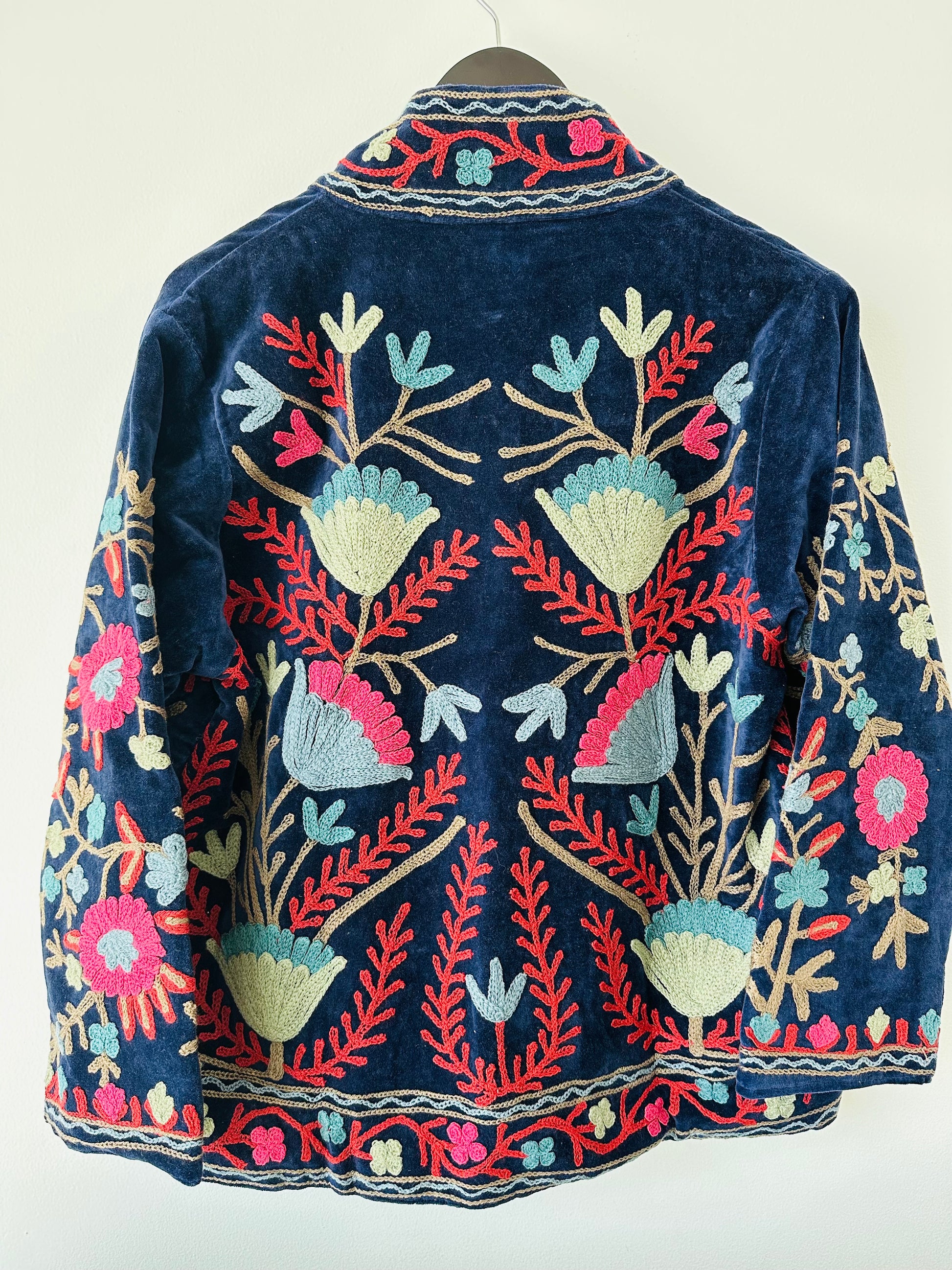 Colorful royal blue embroidered velvet jacket on a hanger against a white wall. Luxurious holiday outfit made by artisans in India for Ornate Handicrafts