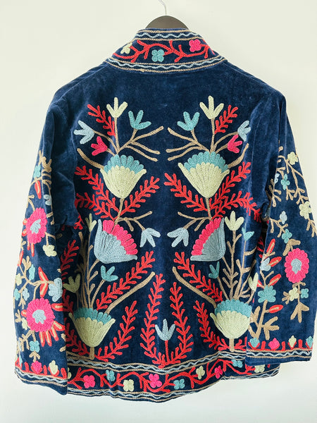 Colorful royal blue embroidered velvet jacket on a hanger against a white wall. Luxurious holiday outfit made by artisans in India for Ornate Handicrafts